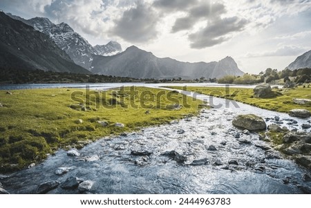 Blue mountain lake and river in a mountain valley among vegetation against the backdrop of rocky peaks with snow in the Fan Mountains in Tajikistan, Tien Shan highlands in the evening