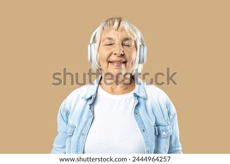 Cheerful elderly blonde woman lady 60s years old in casual clothes listening music with headphones keeping eyes closed isolated on pastel beige background Royalty-Free Stock Photo #2444964257