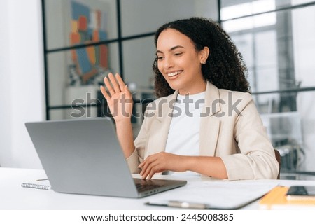 Online video conference. Brazilian or hispanic business woman, corporate manager, team leader, sits in the office, looks at laptop screen, makes greeting sign, talking with colleagues by video call Royalty-Free Stock Photo #2445008385