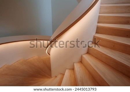 Interior staircase featuring a smooth curve, with warm light emanating from the built-in illumination along the handrail.  Royalty-Free Stock Photo #2445013793