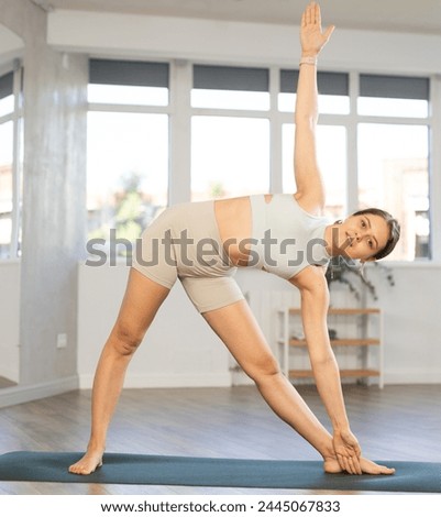 Positive young woman performing forward bending Triangle Pose Trikonasana with extended arms, enhancing flexibility and balance during daily yoga practice in fitness studio Royalty-Free Stock Photo #2445067833