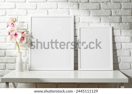 Two Blank Photo Frames on a White Table Against a Brick Wall With Orchids in Vase