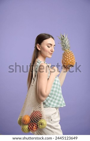 Woman with string bag of fresh fruits holding pineapple on violet background