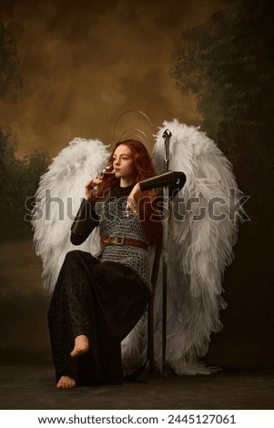 Portrait of young redhaired woman drinking wine while sitting dressed like warrior and holds sword against vintage studio background. Concept of historical and modern times, beauty and fashion. Ad