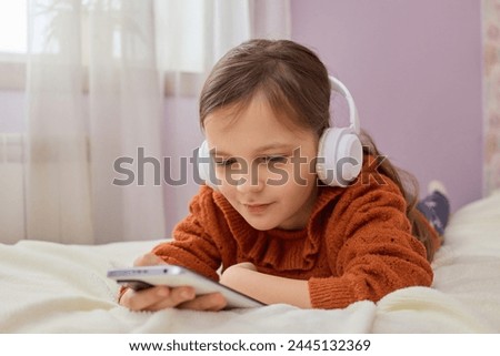 Kid's internet content. Caucasian little girl playing on smartphone using mobile app listening music with headset and watching cartoons lying on her bed at home