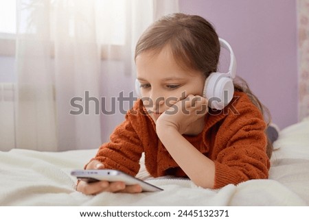 Brown haired little girl in children's room with headphones and smartphone using mobile phone with headset and watching videos at home scrolling online