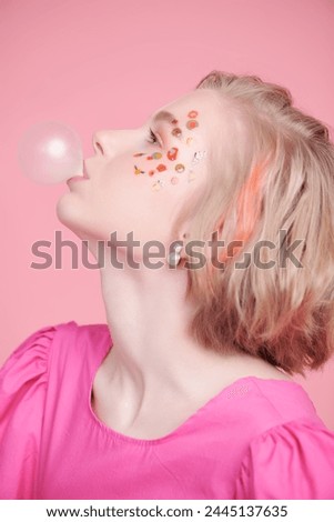 A cute blonde teenage girl with a short haircut poses in a pink dress and makes a bubble with gum. Pink background. Lovely spring-summer look.