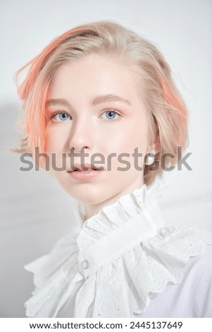 A charming teenage girl with a short haircut and blonde hair with orange streaks poses in a white blouse with a lace collar on a white background. Beauty, style and fashion.