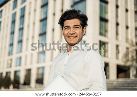 A cheerful young professional in a crisp white shirt holds a laptop, exuding confidence in an urban setting with modern architecture.


