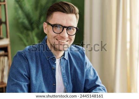 A poised gentleman donning a denim shirt and glasses, his countenance alight with a subtle, congenial smile, positioned against an indoor backdrop with verdure.

 Royalty-Free Stock Photo #2445148161