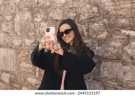 Happy young woman photographing herself using her mobile phone. Female taking selfie with her smart phone at city. Lady with brunette hair making photo outside wear black glasses.