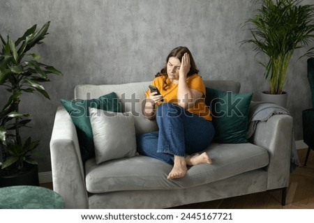 Portrait of beautiful young woman with depressed facial expression sitting on the couch holding her phone. Sad 30s women desperately looking at smartphone screen.