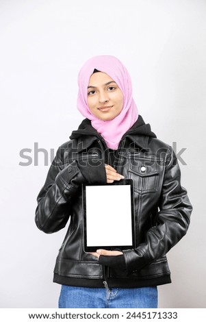 Copy space on her tablet. Confident young Pakistani Muslim woman pointing her digital tablet and smiling while standing, wearing Pink Hijab and a leather jacket isolated on white