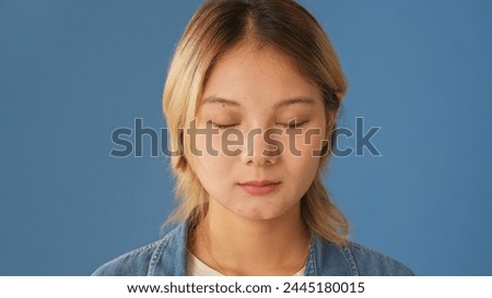 Young woman dressed in denim shirt stands with her eyes closed isolated on blue background in studio