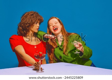 Young redhead woman drinking cocktail and talking to mannequin against blue background. Friends meeting, talking rumors, laughing. Concept of pop art photography, creativity