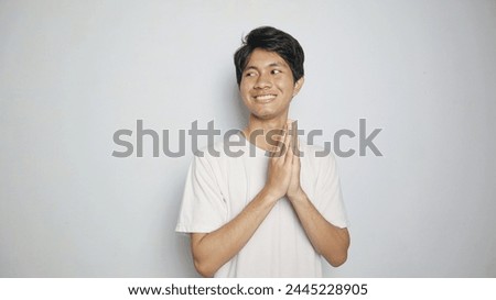 smiling young asian man in white shirt making traditional greeting gesture on isolated white background