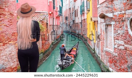 A stylish blonde woman on the bridge watching the Venetian gondolier - Venetian gondolier punting gondola through green canal waters of Venice Italy Royalty-Free Stock Photo #2445229631