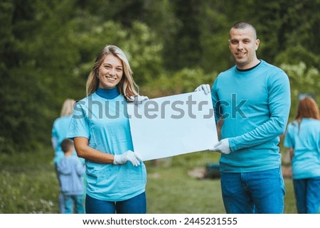 A couple holding a poster in their hands and showing it to the camera, posing standing in nature, smiling at the camera.