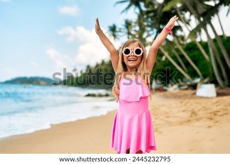 Happy little girl in pink dress, sunglasses smiles, arms raised in joy on sandy tropical beach, with clear blue sky, coconut palms, playful atmosphere in sunny daylight for family vacation memories. Royalty-Free Stock Photo #2445232795
