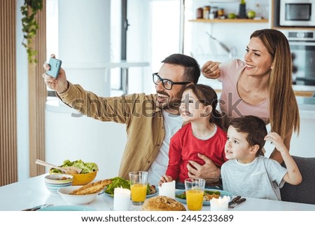 Smiling young family with small preschool children sitting at a table in the kitchen, taking a self-portrait on the phone together, happy parents with small children having fun and taking selfies 