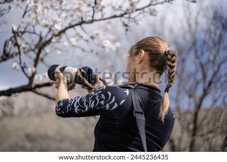 Rear view of young woman takes photos on digital camera in early spring park, the girl taking pictures of blossoming tree, outdoor photographer, travel journey reportage
