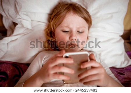 A little girl is lying in bed and using a mobile phone.