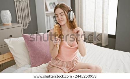 A young woman listens to music with headphones and brushes her hair while sitting on a bed in a cozy bedroom interior.