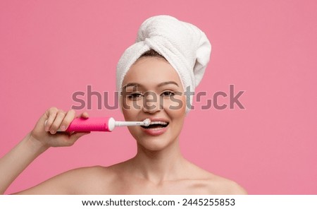 attractive young smiling woman in white bath towel posing on pink background isolated after shower with electric tooth brush, skin care procedures brushing teeth, happy colorful mood