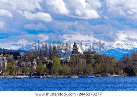 Lakeshore of Lake Zürich with trees and snow covered Swiss Alps in the background at Swiss City of Zürich on a blue cloudy spring day. Photo taken April 2nd, 2024, Zurich, Switzerland.