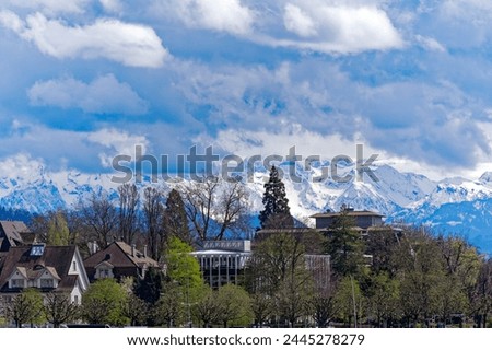 Lakeshore of Lake Zürich with trees and snow covered Swiss Alps in the background at Swiss City of Zürich on a blue cloudy spring day. Photo taken April 2nd, 2024, Zurich, Switzerland.