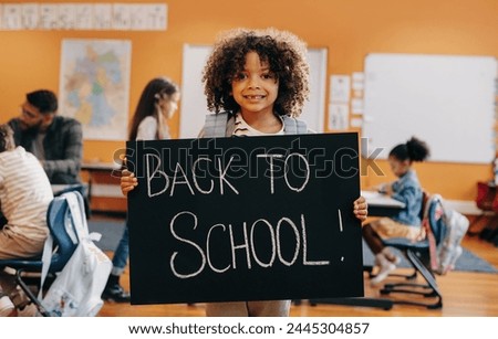 Proud pupil standing in front of co-ed classroom on first day of elementary school, holding a back to school sign. Boy excited to start a new class with his fellow students.