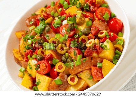 spanish potato salad of chorizo, green olives, cherry tomatoes, red pepper and cilantro with honey mustard dressing in white bowl on wooden table, close-up, dutch angle view Royalty-Free Stock Photo #2445370629