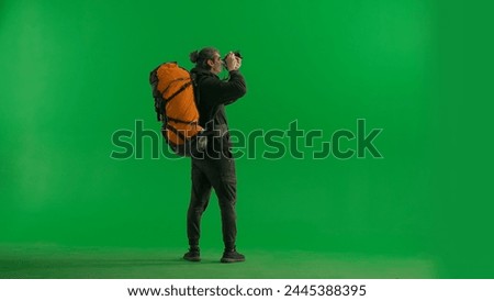 A male traveler takes pictures with a camera while hiking. A tourist with a backpack on his back stands full length in a studio on a green screen. Concept of travel, active rest, hiking.