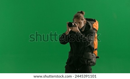 A male traveler takes pictures with a camera while hiking. A tourist with a backpack on his back stands in a studio on a green screen. Concept of travel, active rest, hiking.