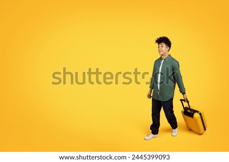 A young black man strolls holding a yellow suitcase, looking at copy space with curiosity against yellow background