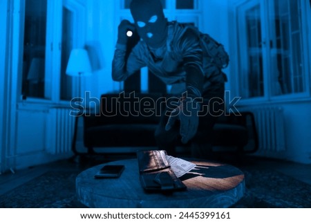 A masked burglar in a dark room uses a flashlight to find and steal a wallet and phone on a wooden table Royalty-Free Stock Photo #2445399161