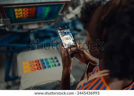 Close up of multicultural printing shop worker's hands taking pictures with cellphone of her finished product.Cropped picture of interracial female printer's hands capturing silk screen printed shirt