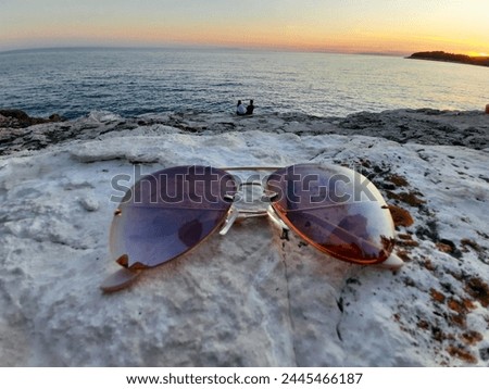 Holidaymood - Sunglases laying on a rock - couple in the background with seaview while sunset Royalty-Free Stock Photo #2445466187
