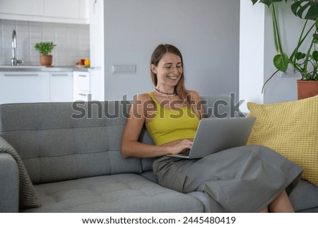 Pleased woman smiling using laptop on knees sitting at couch. Interested female with computer on lap have fun online enjoying comfort, chatting with friends on social media networks, watching video. Royalty-Free Stock Photo #2445480419