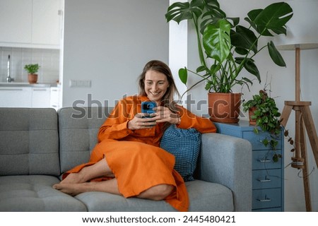 Happy woman using phone on sofa at home. Cheerful female smiling having fun, relaxing, enjoying comfort, chatting with friends on social networks, watch movies, series, play online games on smartphone Royalty-Free Stock Photo #2445480421