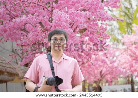 Asian middle-aged man in pink shirt and eyeglasses standing and holding camera in rest area of private park while taking weekend vacation, soft focus, happiness of people concept.