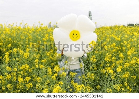 Young woman holds daisy balloon with a smiling face in front of own face in the middle of rapeseed (Brassica napus) field full of bright colourful yellow flowers. Spring, summer, happy, joy concept