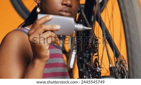 BIPOC technician using specialized lubricant to repair squeaky bicycle chain, orange studio background. Specialist applying oil on noisy bike parts during maintenance process, close up shot Royalty-Free Stock Photo #2445545749