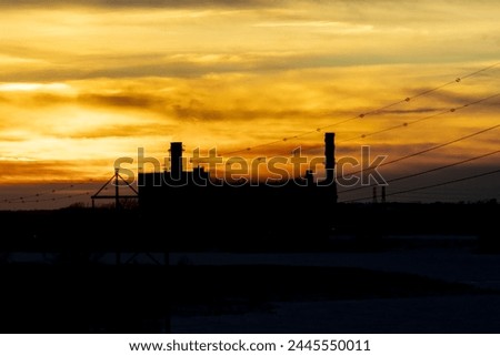 Sunsetting over industrial facility. Silhouette of large refinery with tall exhaust towers.  Power lines reach to the factory. Burning orange sky makes the background