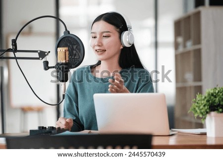 A woman is sitting at a desk with a laptop and a microphone. She is wearing headphones and she is recording a podcast or a voiceover