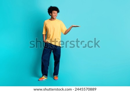 Full size photo of good mood man wear oversize t-shirt denim pants look at object on arm empty space isolated on teal color background