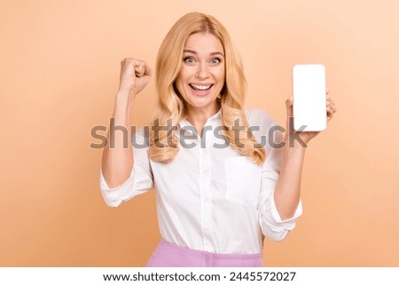 Photo of lucky excited woman wear white shirt rising fist showing modern gadget empty space isolated beige color background
