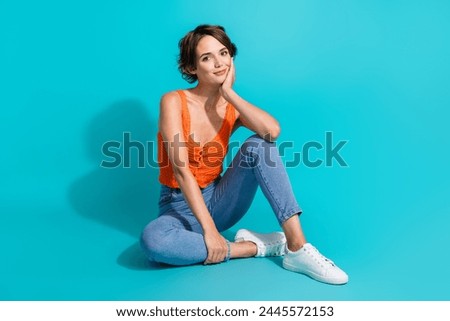 Full length photo of lovely positive woman wear orange singlet jeans sitting on floor palm on cheek isolated on turquoise color background
