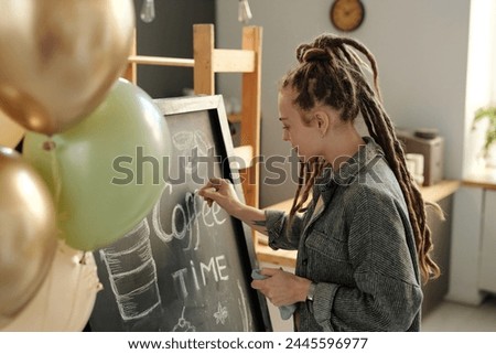 Young businesswoman with dreadlocks standing in front of blackboard and drawing advert of new cafe while preparing for opening after refit Royalty-Free Stock Photo #2445596977