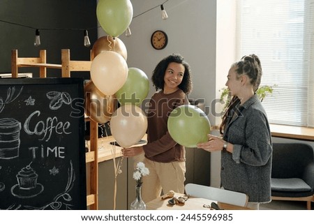 Happy young African American woman with bunch of balloons looking at colleague with dreadlocks while both decorating cafe after refit Royalty-Free Stock Photo #2445596985